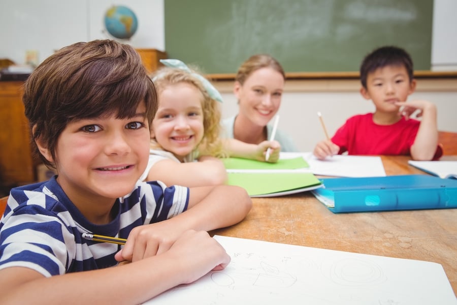 Pretty teacher helping pupil in classroom smiling at camera at the elementary school